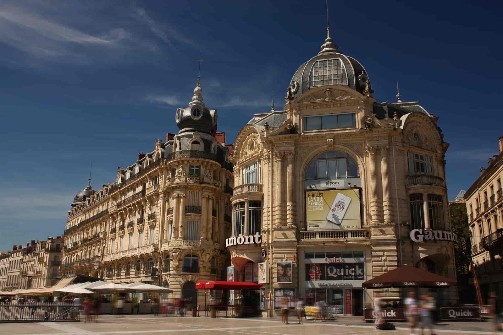 A photo of Place de la Comédie, a famous square in Montpellier, France. The square is filled with people, and there are many buildings around it