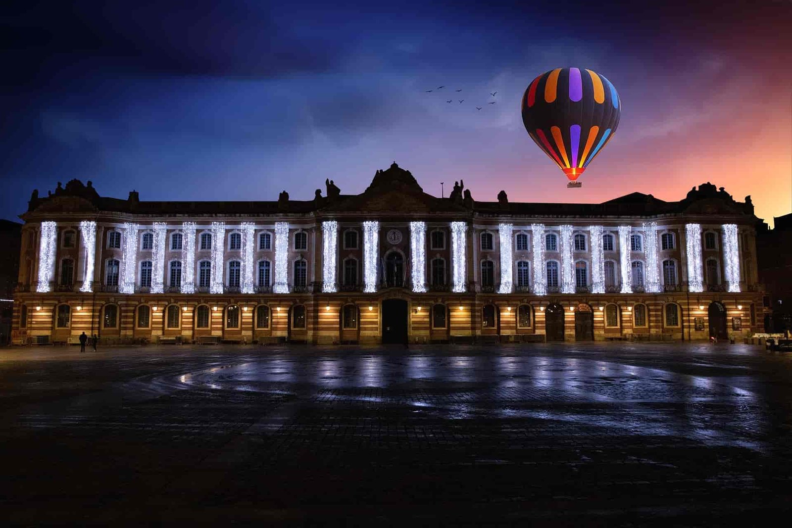 A photo of Place du Capitole, Toulouse, France, at night. The building is lit up with lights, and a hot air balloon is flying above