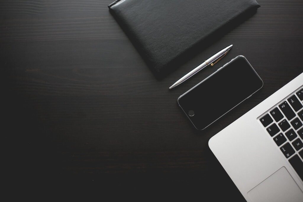 A laptop, notebook, pen, and smartphone arranged on a dark wooden desk